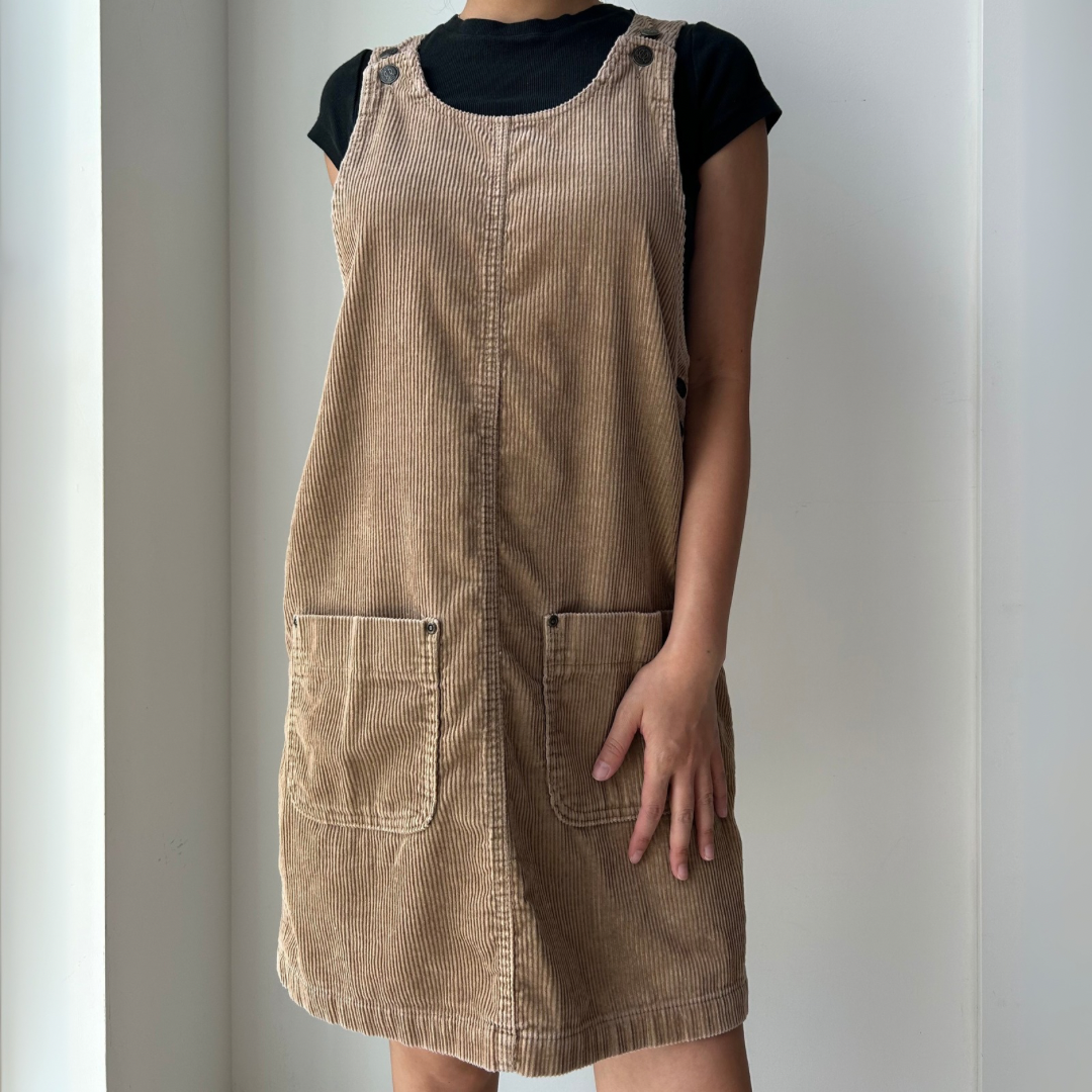 Person wearing a brown corduroy pinafore dress over a black top against a plain background